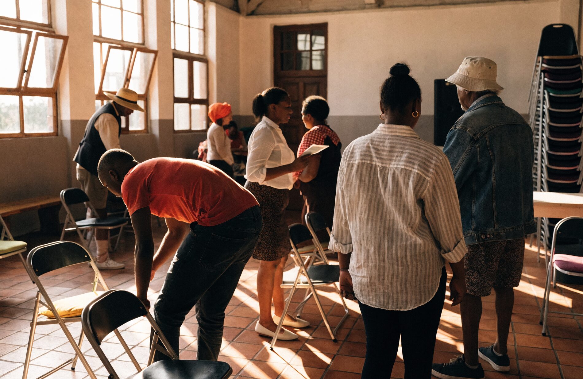 A congregation talks in small groups and sets out folding chairs in a sunlit church hall after a gathering